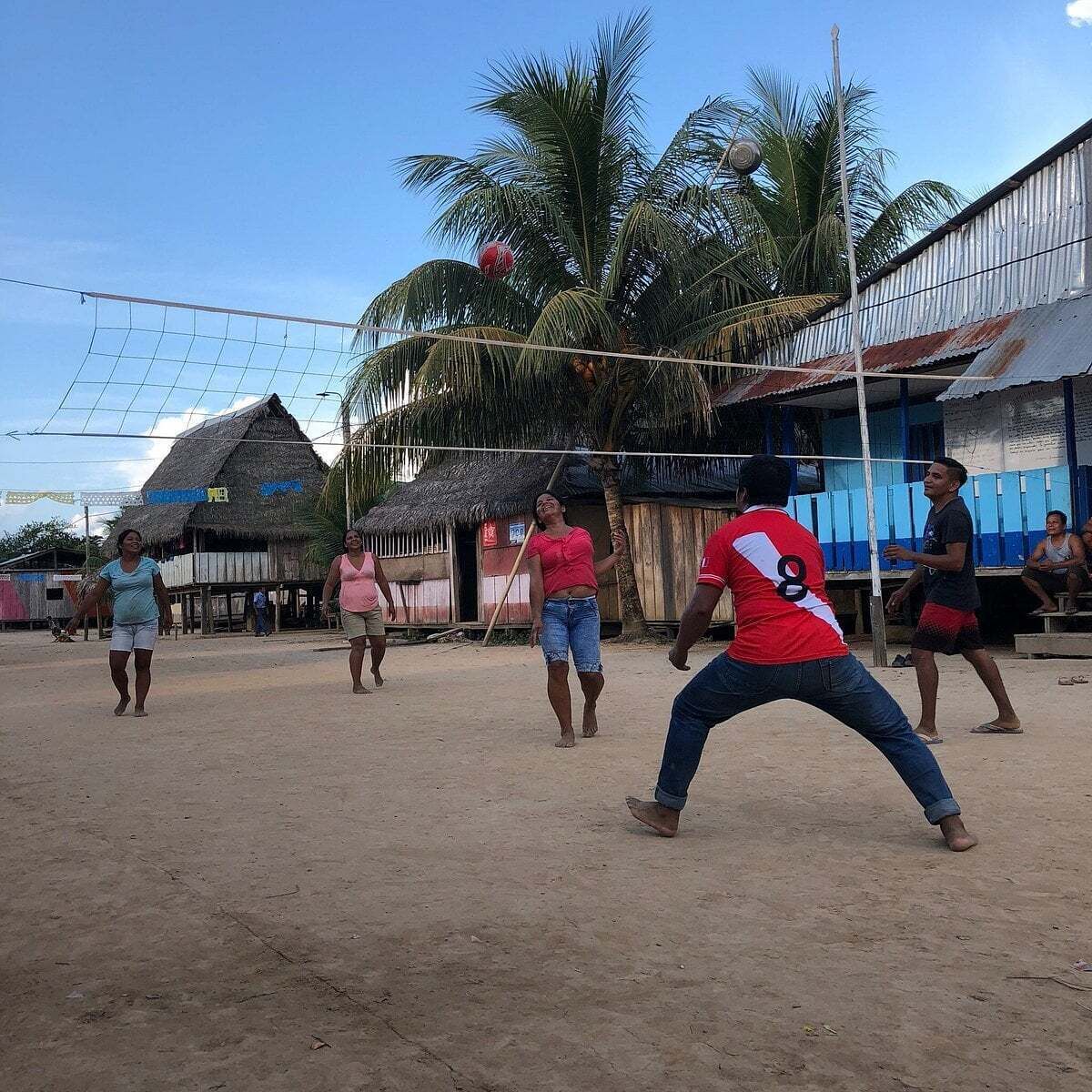 A group of people playing volleyball on a sandy court with thatched-roof buildings in the background.