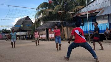 A group of people playing volleyball on a sandy court with thatched-roof buildings in the background.