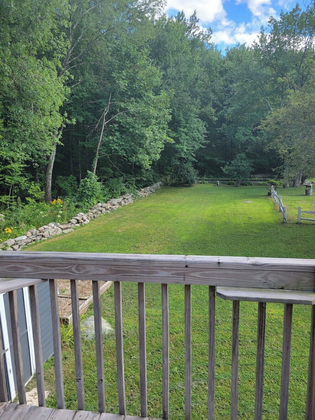 Une terrasse en bois, un mur en pierre et une pelouse entourée d’arbres.