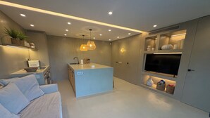 Una sala de estar moderna con sofá, una pequeña cocina, una isla central, iluminación empotrada y un televisor de pantalla plana montado en la pared.