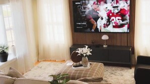 Ein Wohnzimmer mit einer Couch, einem Couchtisch mit einer Vase, einem an der Wand montierten Fernseher und einem Fenster mit Vorhängen.