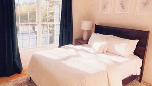 Ein Schlafzimmer mit einem großen Bett, einem Nachttisch mit Lampe und einem Fenster mit Vorhängen.