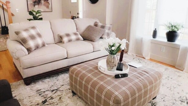 Ein Wohnzimmer mit einem beigen Sofa, einem karierten Ottomanen und einer Blumenvase.