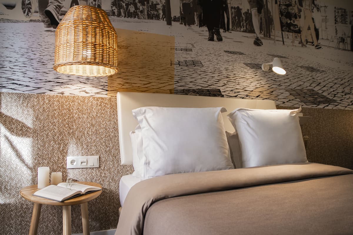 A bed with white bedding, a wicker lamp, a small wooden table with a book, and a wall with a patterned design.