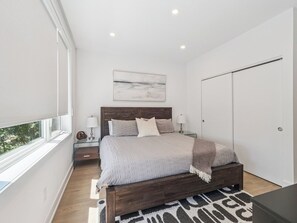A modern bedroom with a large bed, bedside tables, a nightstand, a window with blinds, and a wardrobe.