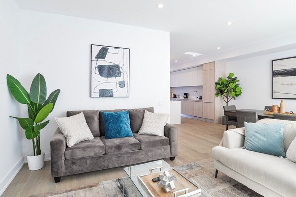A modern living room with a grey sofa, a white sofa, and a coffee table with a remote control. There is a kitchen in the background with white cabinets and a dining area.