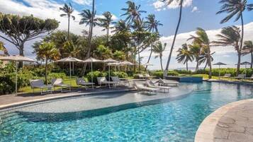A clear blue pool with lounge chairs and umbrellas, surrounded by palm trees and greenery.