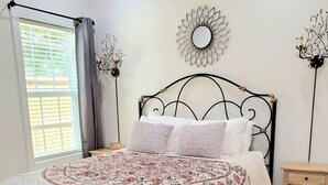 A bedroom with a bed, a bedside table, a decorative mirror, and a window with curtains. - Ruby's Cottage / Close to Beaches & Downtown (Mount Pleasant)