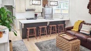 A modern kitchen with white cabinets, a central island with stools, and a dining area with a brown leather couch and a woven coffee table. - Ruby's Cottage / Close to Beaches & Downtown (Mount Pleasant)