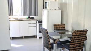 Et kompakt køkken med mikrobølgeovn, et lille køleskab og et spiseområde med et glastop bord og fletstole.