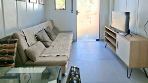 En stue med en beige sofa, et glasbord, et fjernsyn på et træstel og billeder i rammer på væggen.