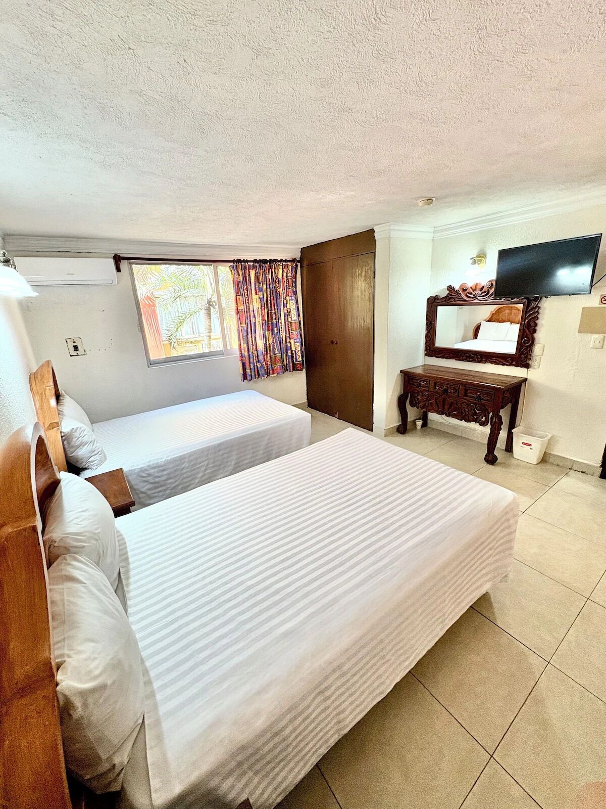 Habitación de hotel con dos camas, televisión, espejo y ventana con cortinas.