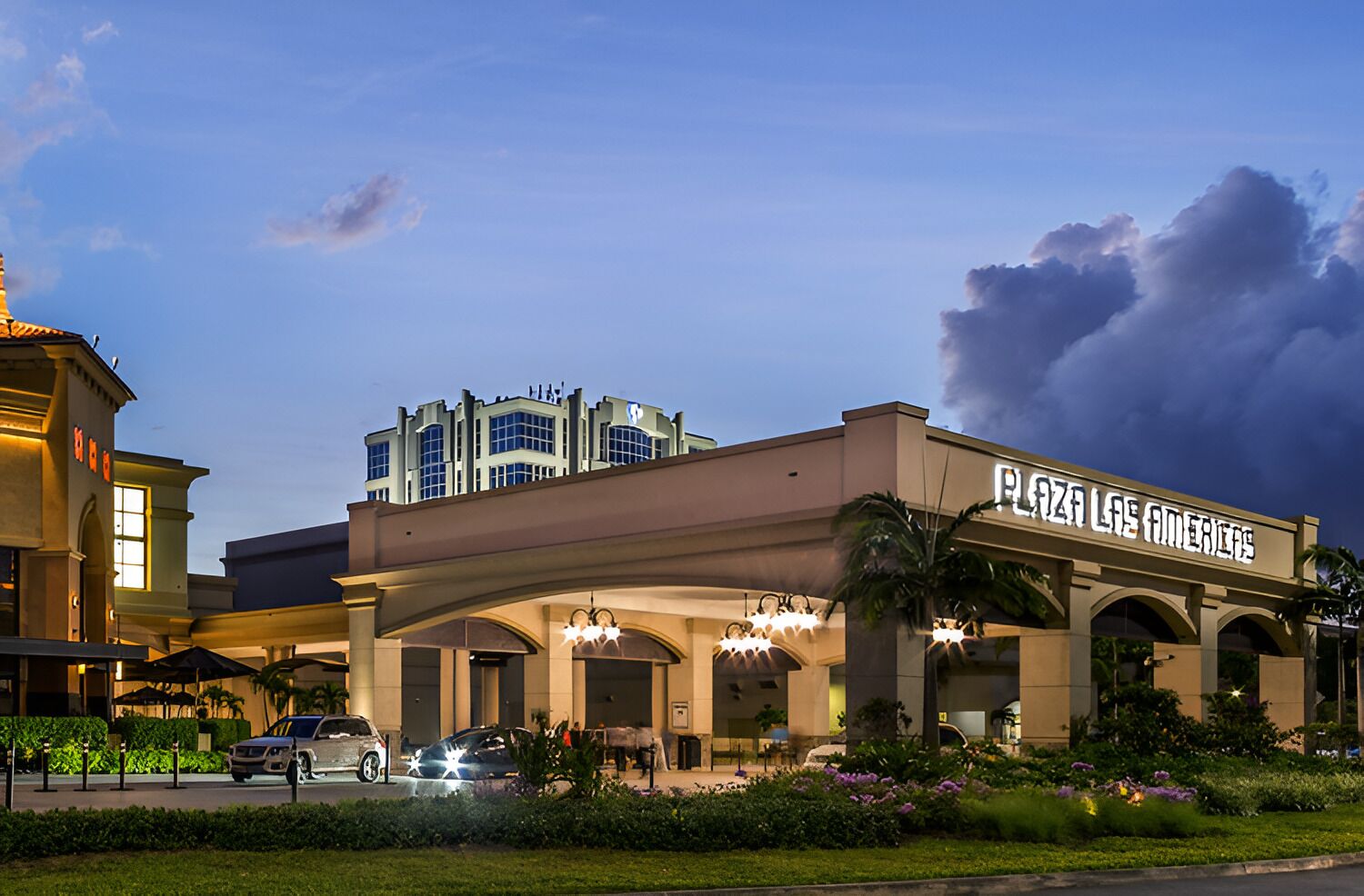 Et hotel med et tydeligt skilt, der viser "Plaza Las Americas". Arkitekturen er moderne med et fladt tag og store vinduer. Hotellet er omgivet af velplejede beplantninger og har en parkeringsplads foran.