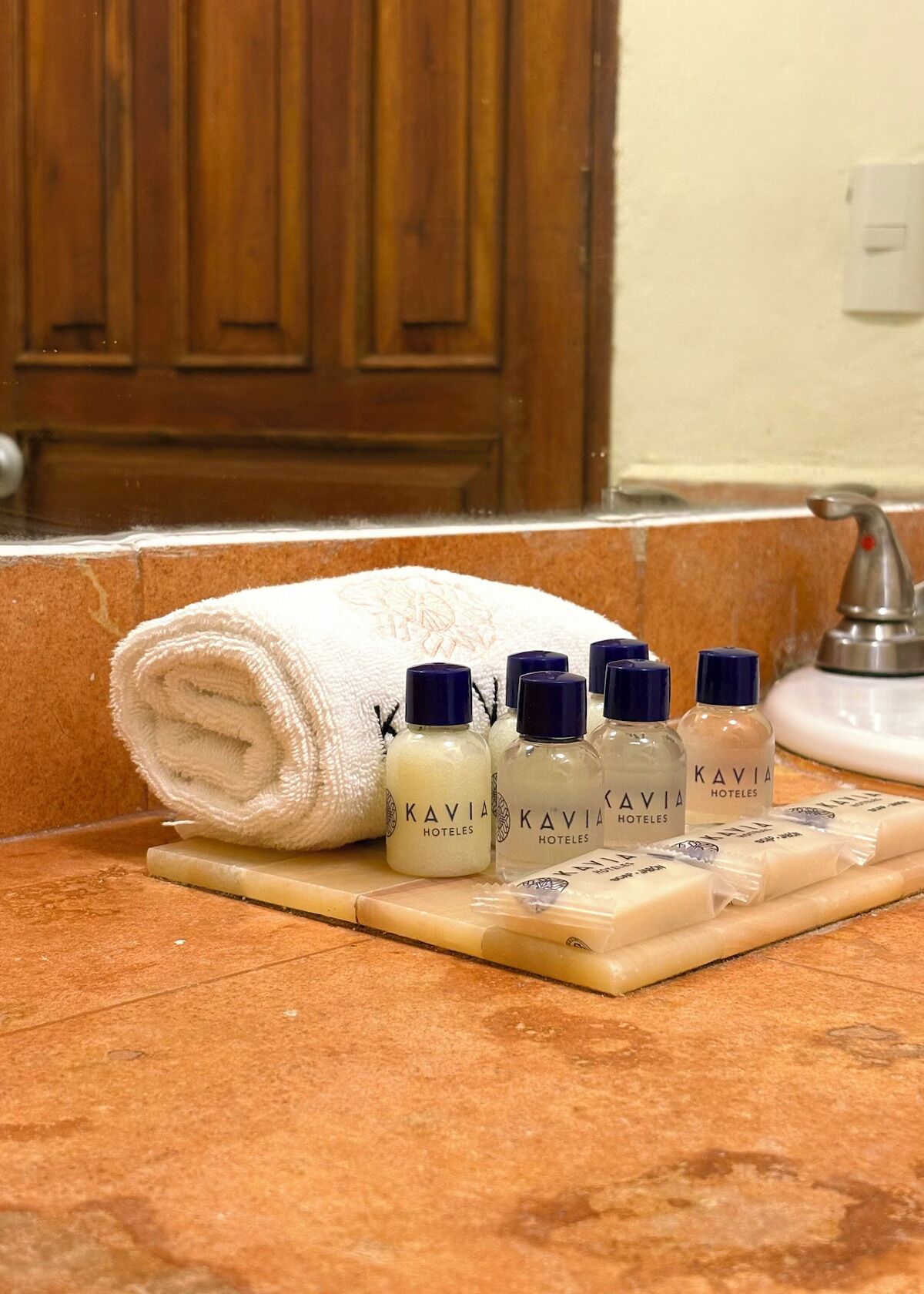 A bathroom counter with a rolled towel, several bottles of Kavia Hotels toiletries, and a soap bar.
