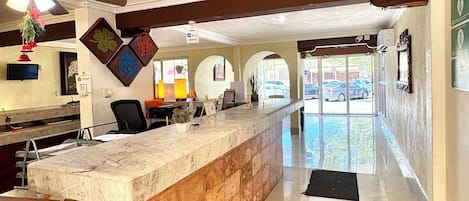 Zona de recepción de un hotel con mostrador de mármol, ventilador de techo y vista a coches estacionados afuera.