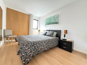 Un dormitorio con una cama, una silla, una mesita de noche, una lámpara y una ventana.