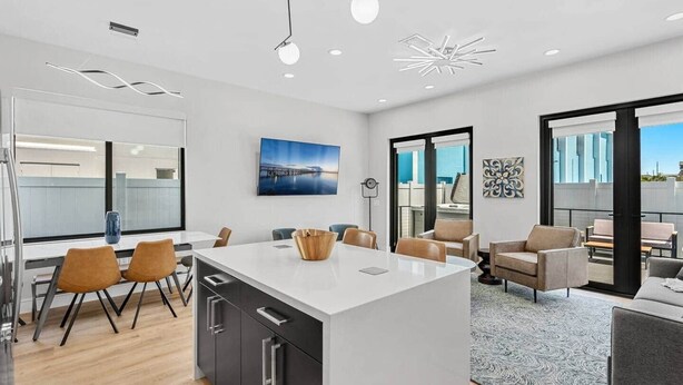 A modern kitchen with a central island, dining area with chairs, and a view of the cityscape through large windows. - Across The Beach • 4 Units • Sleeps 28 • Hot Tubs (Cocoa Beach)