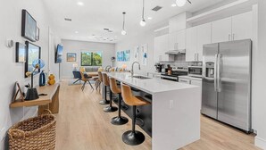 A modern kitchen with a central island, stainless steel appliances, and a dining area with bar stools. - Across The Beach • 4 Units • Sleeps 28 • Hot Tubs (Cocoa Beach)