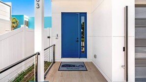 A modern building entrance with a blue door, a welcome mat, and a white fence. - Across The Beach • 4 Units • Sleeps 28 • Hot Tubs (Cocoa Beach)