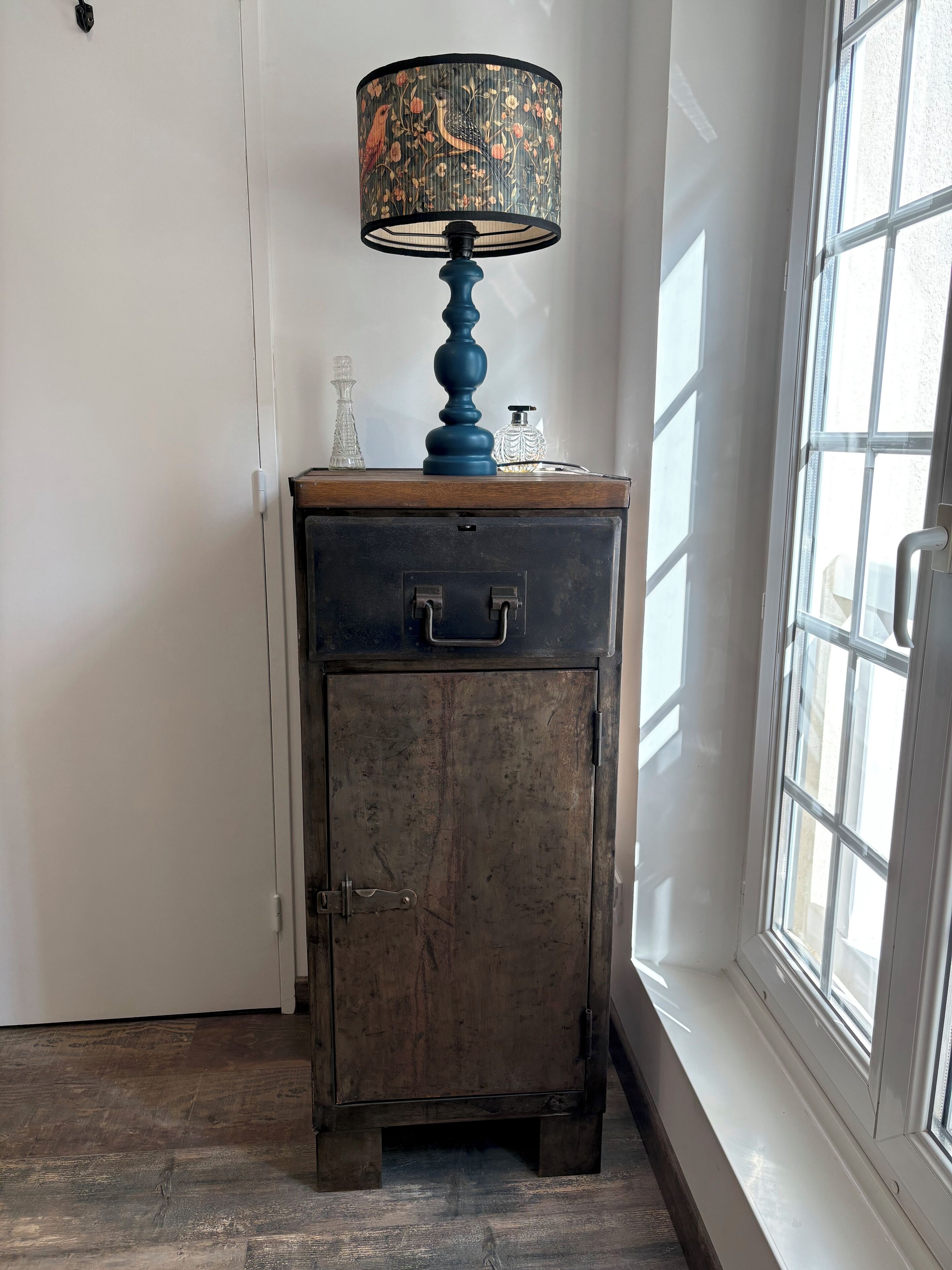 A vintage-style lamp with a floral pattern on a wooden cabinet with a drawer, placed next to a window with a view of the outside.