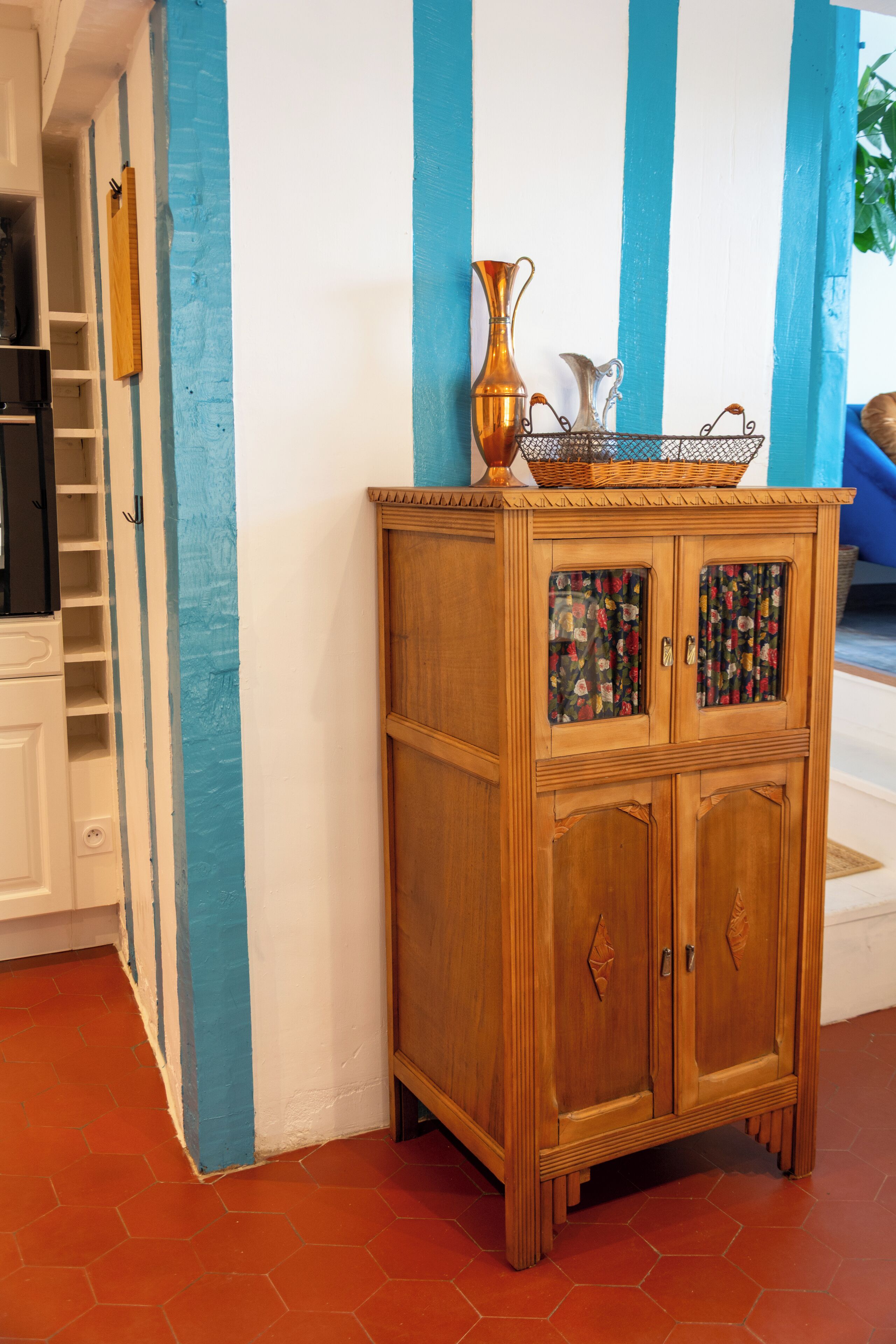 Een houten kast met deuren van glas met bloemmotief, een koperen vaas en een rieten mandje erbovenop.
