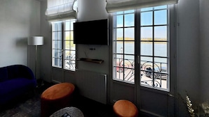 Ein Zimmer mit an der Wand befestigtem Fernseher, zwei Fenstern mit weißen Vorhängen, einem blauen Sofa und zwei orangefarbenen Hocker.