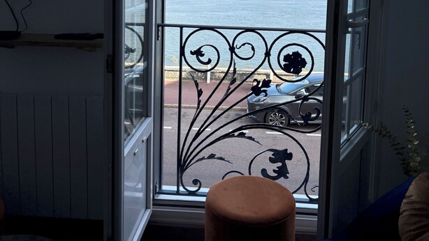 Ein Zimmer mit großem Fenster und Blick auf ein Gewässer sowie eine entfernte Küstenlinie.
