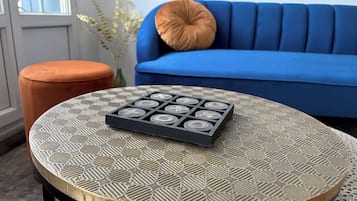 Una mesa redonda con tablero de diseño geométrico, una bandeja negra con piezas de juego, un sofá azul con cojín marrón y un reposapiés naranja.