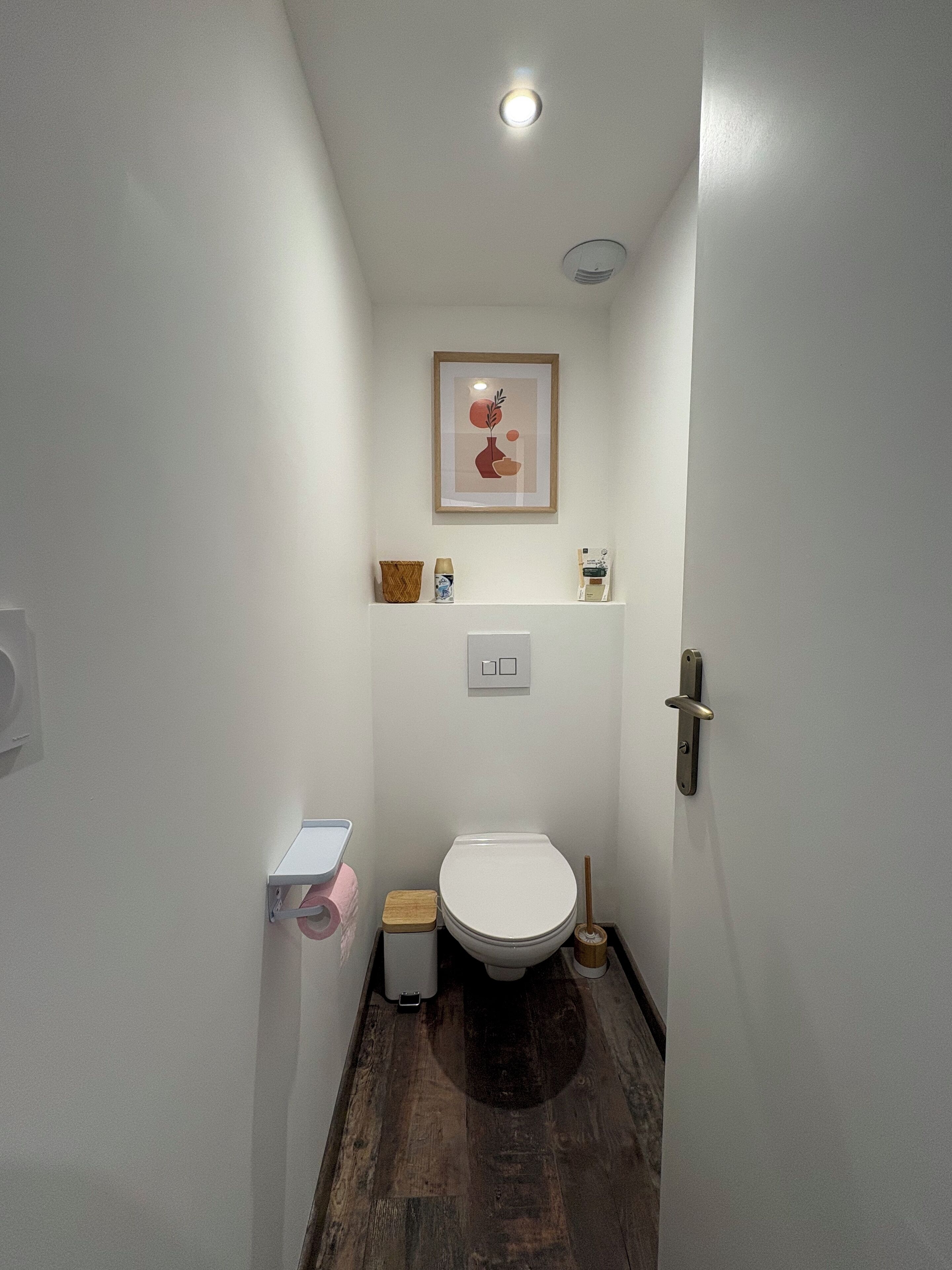 Ein schmales Badezimmer mit Toilette, einer kleinen Ablage für Toilettenartikel, einem gerahmten Bild an der Wand und Holzboden.