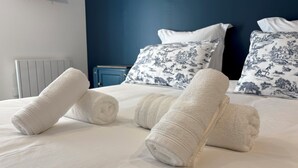Ein ordentlich bezogenes Bett mit aufgerollten weißen Handtüchern und gemusterten Kissen in einem Raum mit blauer Akzentwand und einem weißen Heizkörper.