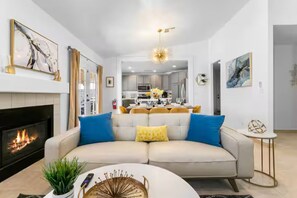A modern living room with a fireplace, a sofa with cushions, a coffee table with decorative items, and a dining area in the background. - 5BD Memorable Stays and Backyard Games Galore! (North Las Vegas)