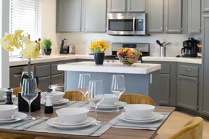 A modern kitchen with grey cabinets, a stainless steel microwave, and a dining area set with white plates, glasses, and a fruit basket. - 5BD Memorable Stays and Backyard Games Galore! (North Las Vegas)