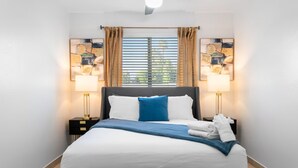 A neatly made bed with white and blue bedding, two bedside lamps, and a window with blinds.