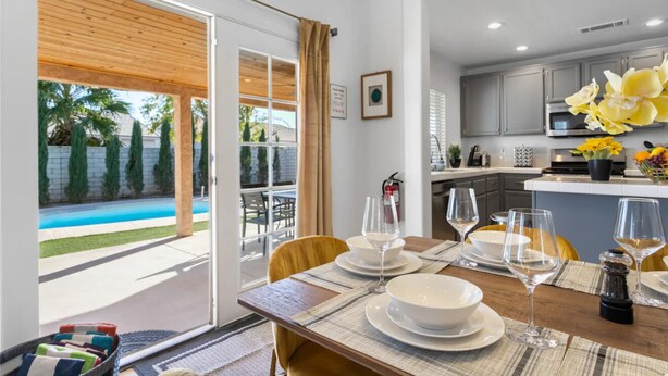 A dining area with a table set for four, a kitchen in the background, and a pool visible through the glass door.