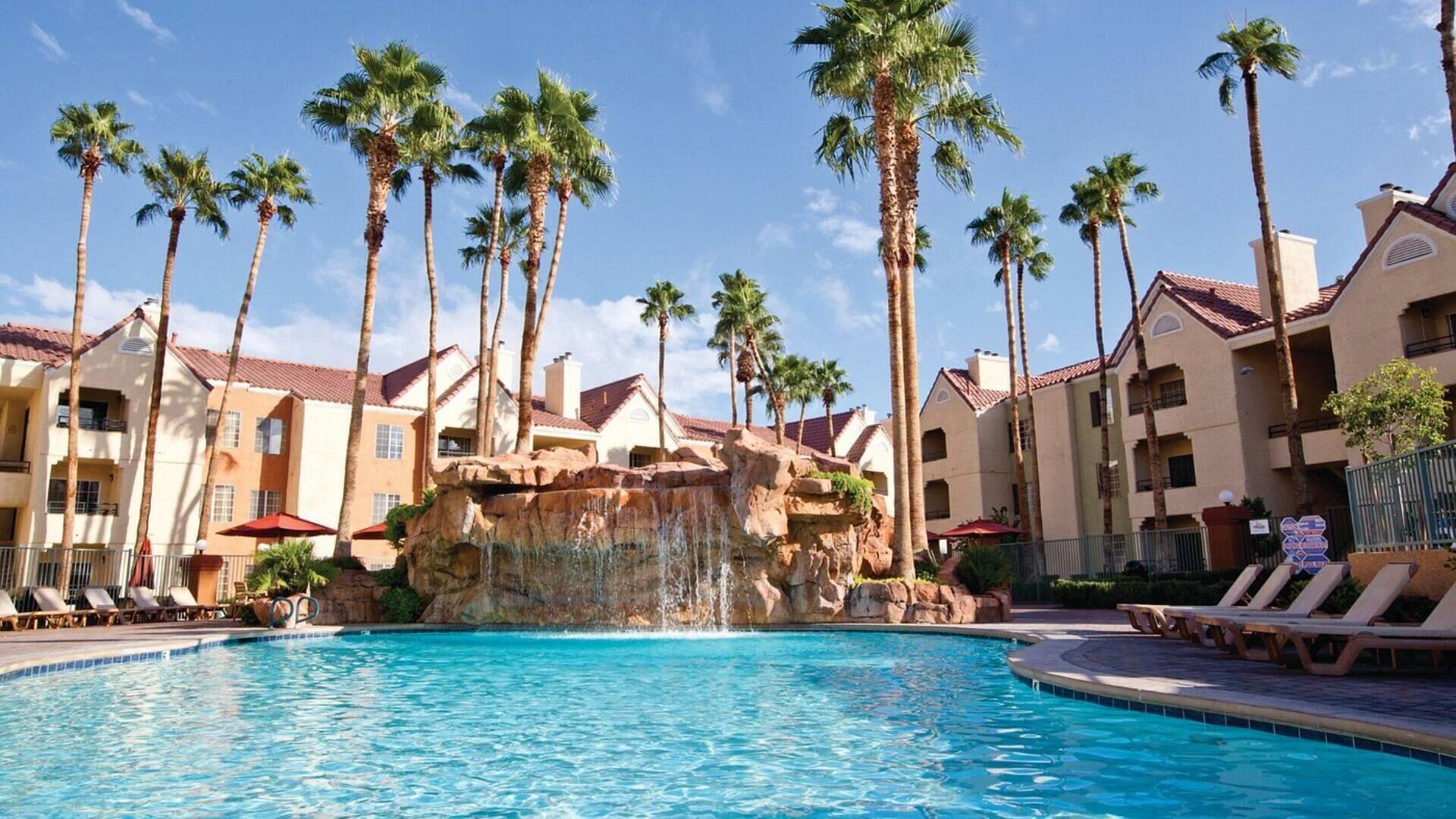 A swimming pool with palm trees and a waterfall feature, surrounded by hotel buildings with balconies and red roofs.