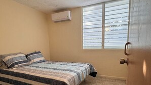 Une chambre à coucher avec un lit, une fenêtre avec des stores, un appareil de climatisation et une porte.