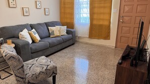 Un salon avec un canapé gris, un pouf à motifs et une table d’appoint en bois sur laquelle se trouvent une télévision et une télécommande.