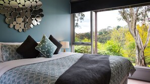 Une chambre à coucher avec un lit, des oreillers, un miroir mural et une fenêtre donnant sur des arbres.