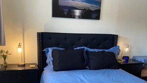 A neatly made bed with a blue bedspread and a quilt, flanked by two bedside tables with lamps, a wall-mounted painting, and a carpeted floor. - Lake Room (Sandpoint)