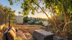 Eine Terrasse mit einem Rattan-Sofa und einer gepolsterten Bank, umgeben von Bäumen und Blick auf eine Wohngegend.