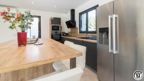 A modern kitchen with a wooden table, white chairs, a stainless steel refrigerator, and a built-in oven.