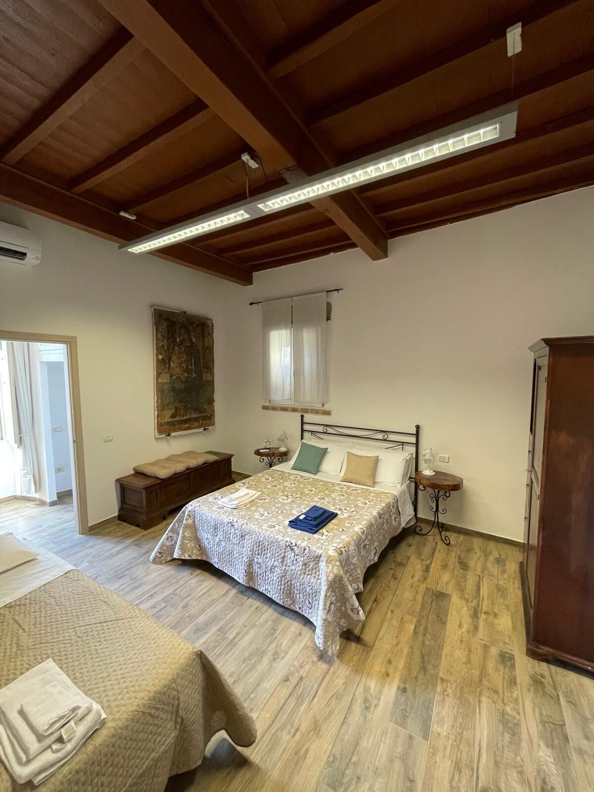 Quarto com teto de madeira, cama com colcha estampada, mesa de cabeceira com abajur e um banco de madeira.