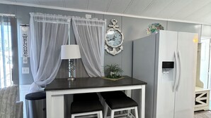 A compact kitchen with a white refrigerator, a small dining table with two chairs, a clock on the wall, and a window with sheer curtains.