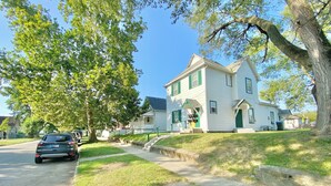 Une maison à deux étages avec une porte d’entrée verte et un revêtement extérieur blanc, une allée et un grand arbre à l’avant.