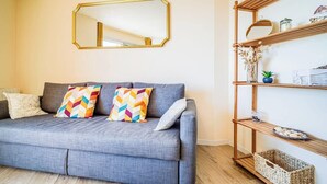 A grey sofa with colorful pillows, a gold-framed mirror, and a wooden shelf with decorative items. - Appartement bord de plage, havre de paix (Ploemeur)