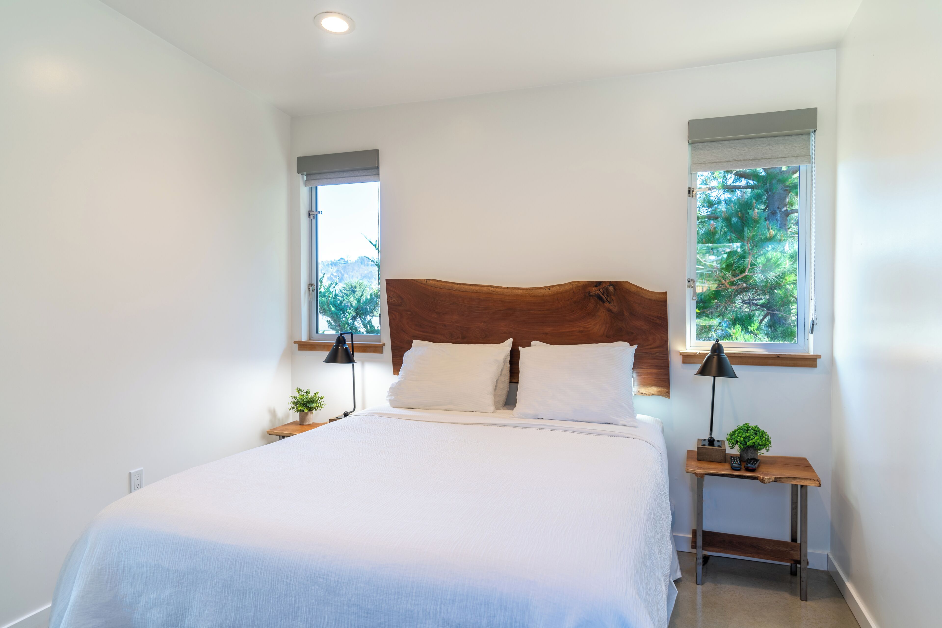 A bedroom with a large bed, two bedside tables, a wooden headboard, and two windows with blinds.