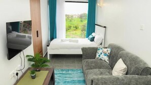 Una habitación compacta con cama, sofá gris, televisor montado en la pared y ventana con cortinas.