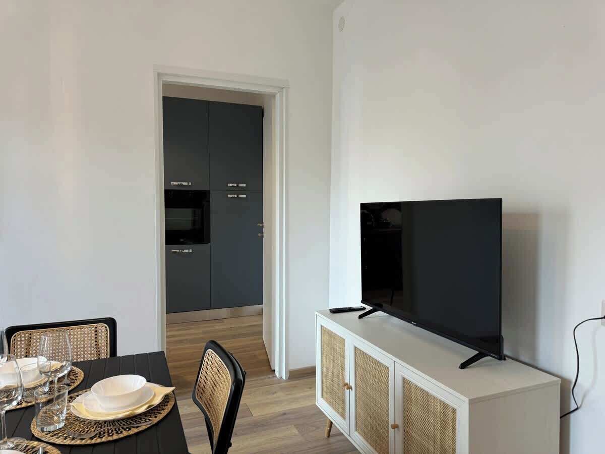 Ein modernes Wohnzimmer mit Essbereich, einem Flachbildfernseher auf einem weißen Schrank und einer Küche im Hintergrund.