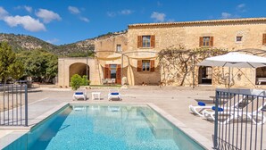 Zona de piscina con sillones de descanso y sombrillas, un edificio de piedra con ventanas y persianas, y un paisaje montañoso al fondo.