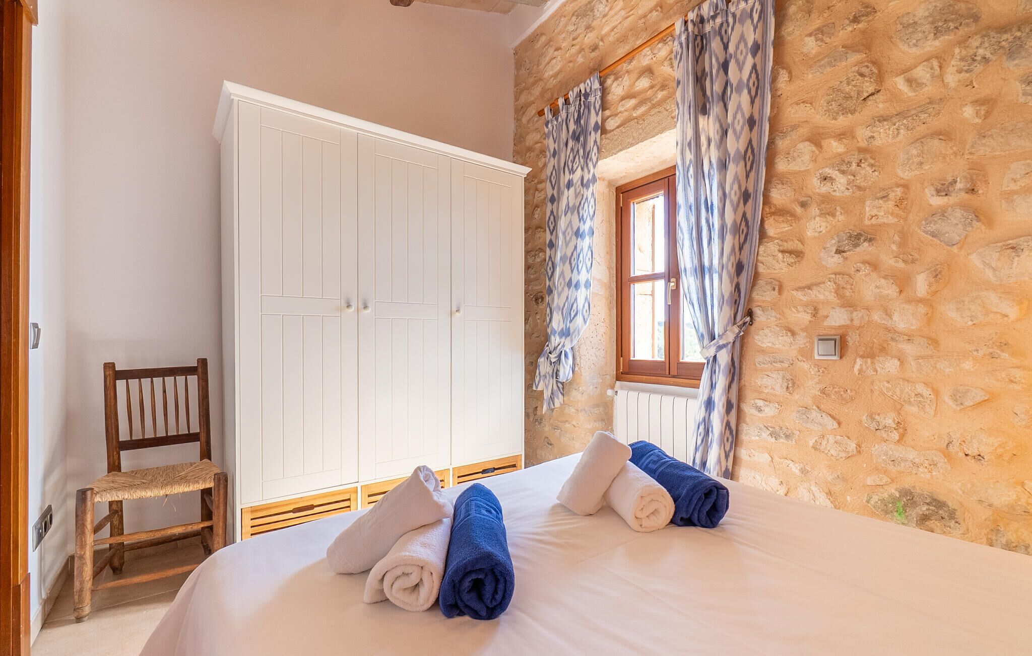 Une chambre avec un mur en pierre, une chaise en bois, une armoire blanche et un lit avec des serviettes.